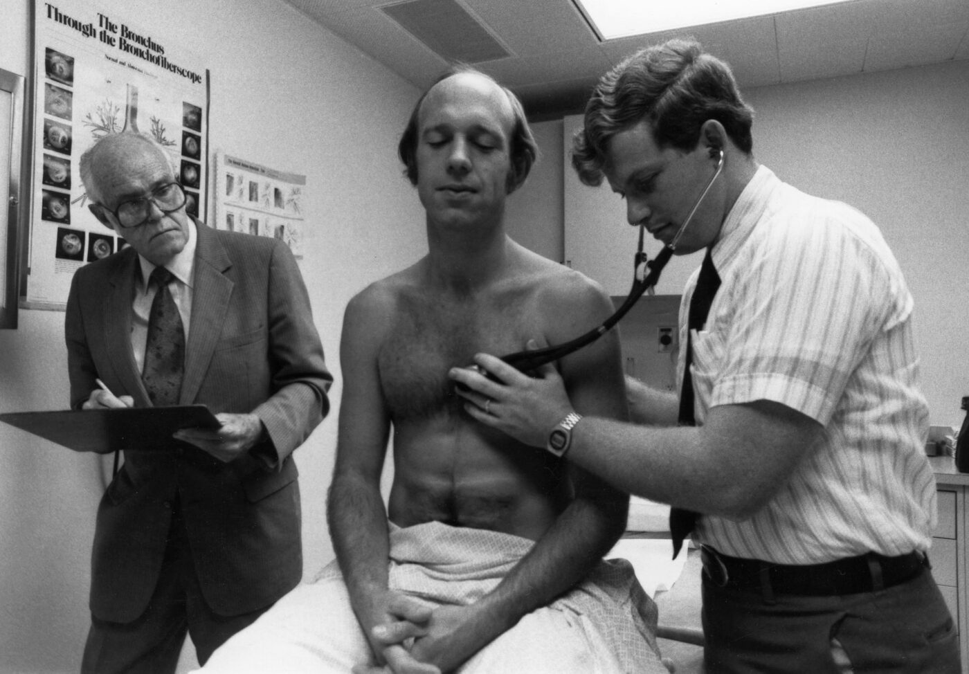 A Kettering College Physician Assistant Studies student (circa 1976) works on another student while a professor takes notes. 